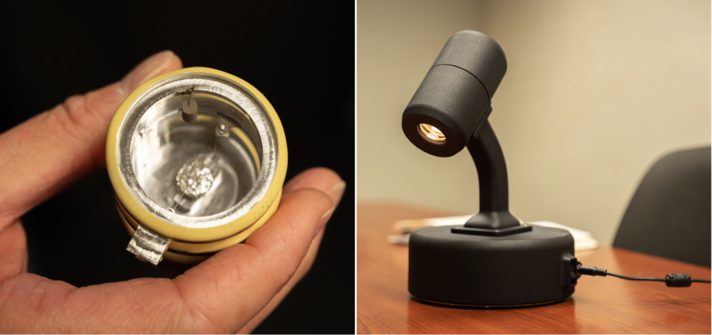 Two side-by-side photos. Left: a hand holds a gold and silver cylindrical device with a glass window, about the size of a light bulb. Right: A sleek black lamp shines down on a brown tabletop.